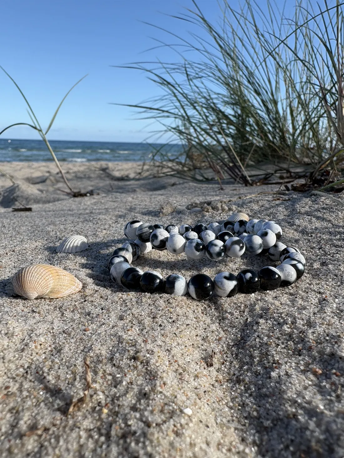 Zwei OSTSEE WAL Spendenarmbänder aus Orca-Achat im Ostseesand, daneben Muscheln und Dünengras